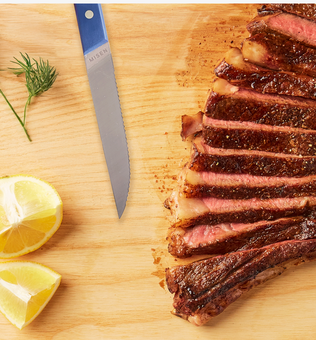Sliced ribeye steak on wooden cutting board with Misen steak knife and lemon wedges
